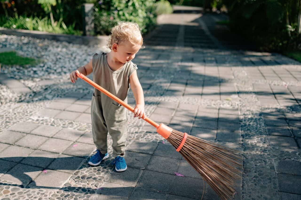charming child sweeping concrete pavement with broomstick